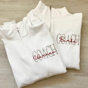 May include: Two white quarter-zip sweatshirts with the word "COACH" embroidered in gray with a name in red underneath. The sweatshirt on the left says "Warren" and the sweatshirt on the right says "Burke".