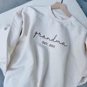May include: A white sweatshirt with the text "grandma est. 2023" embroidered in brown.