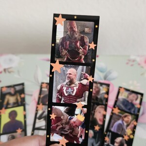 May include: A black bookmark featuring three rectangular images of a man in a red and white costume. The bookmark is decorated with orange star stickers. Other bookmarks with similar images are visible.