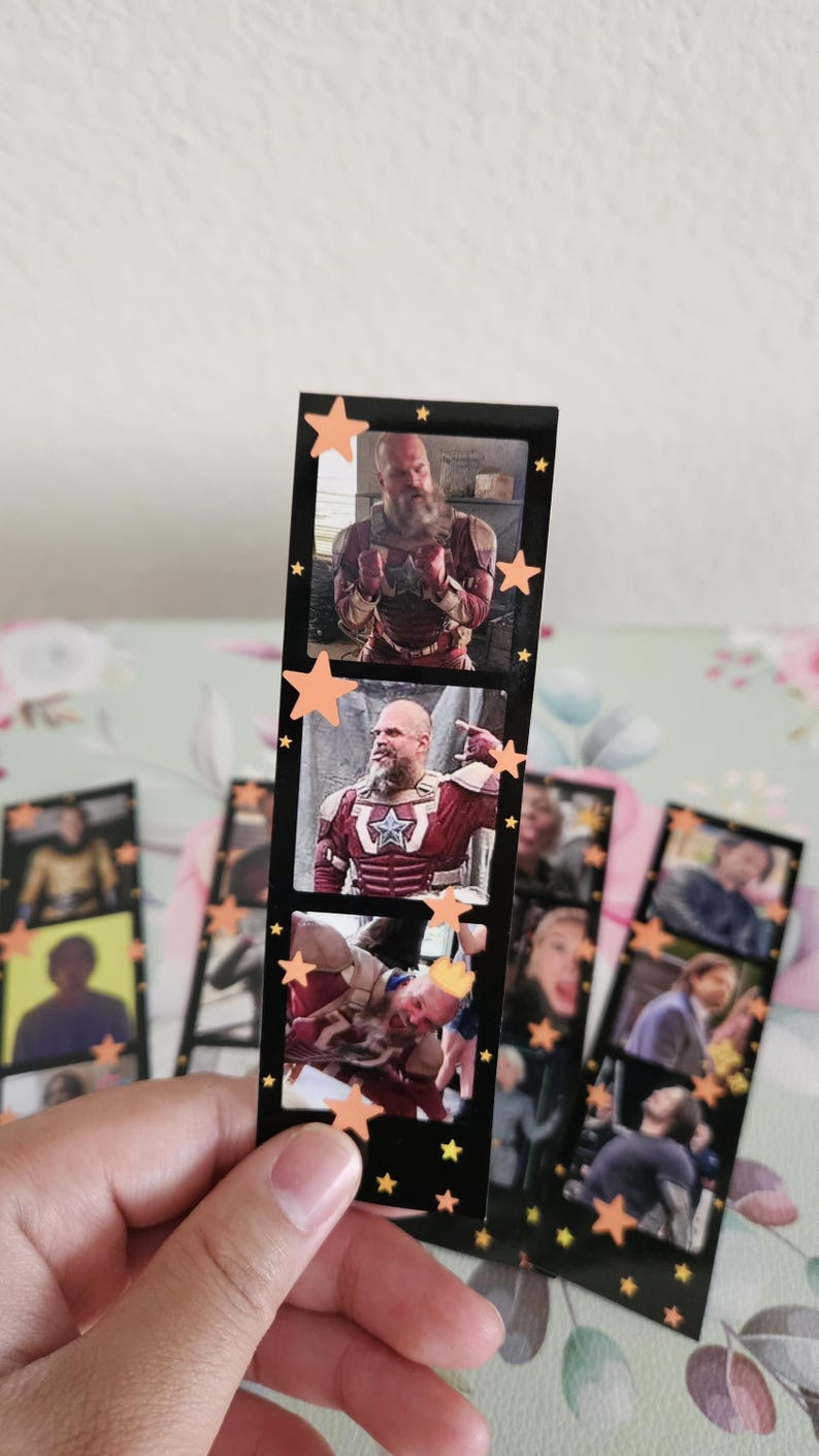 May include: A black bookmark featuring three rectangular images of a man in a red and white costume. The bookmark is decorated with orange star stickers. Other bookmarks with similar images are visible.