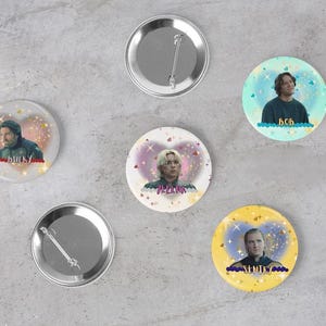 May include: A collection of six round metal pinback buttons. Five buttons feature images of people with text, set against colorful backgrounds. Two buttons are blank, showing the silver backs and pin fasteners.