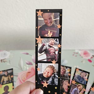 May include: A black film strip featuring three images of a smiling man in a costume, with orange star accents. The film strip is held in a hand, with other similar strips visible in the background. The images are of a man in a costume.