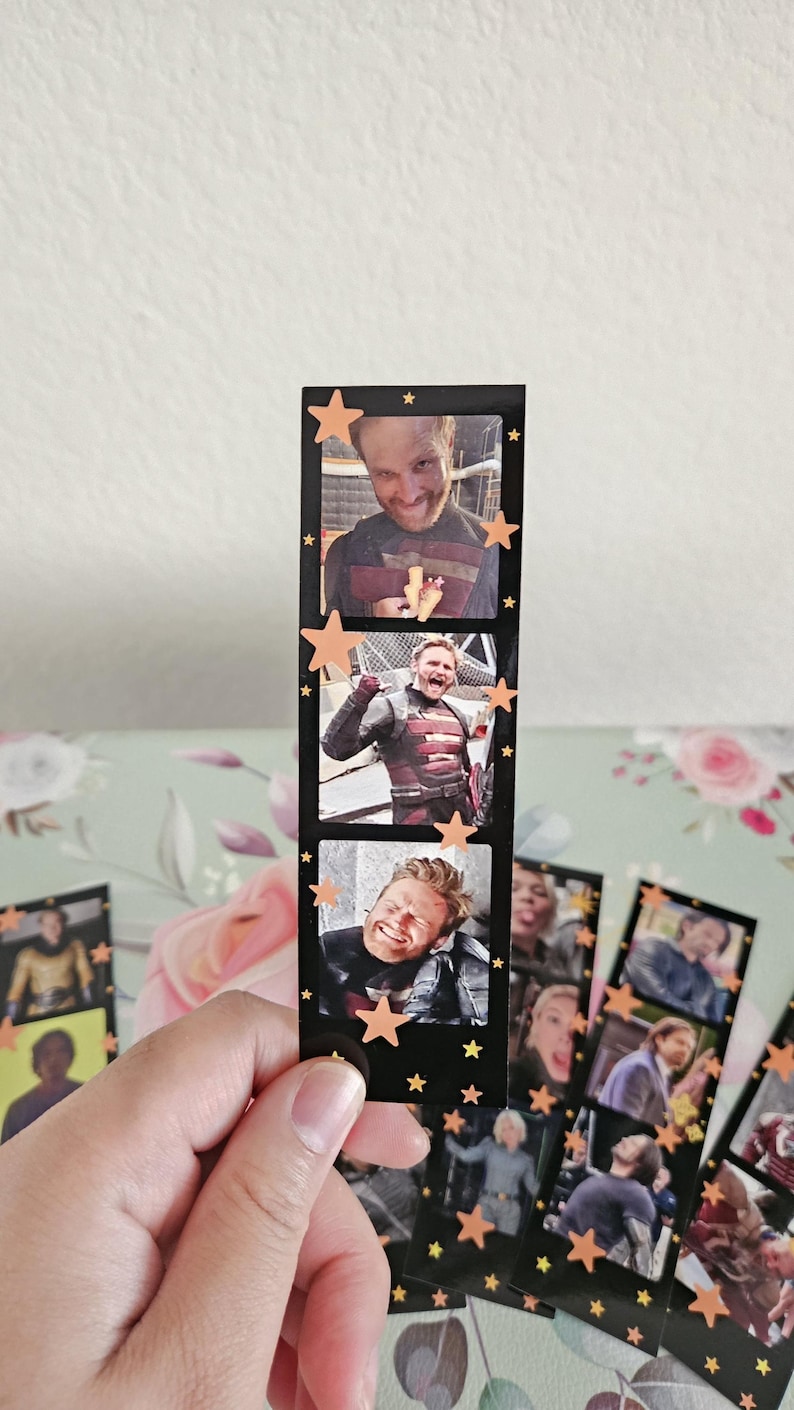 May include: A black film strip featuring three images of a smiling man in a costume, with orange star accents. The film strip is held in a hand, with other similar strips visible in the background. The images are of a man in a costume.