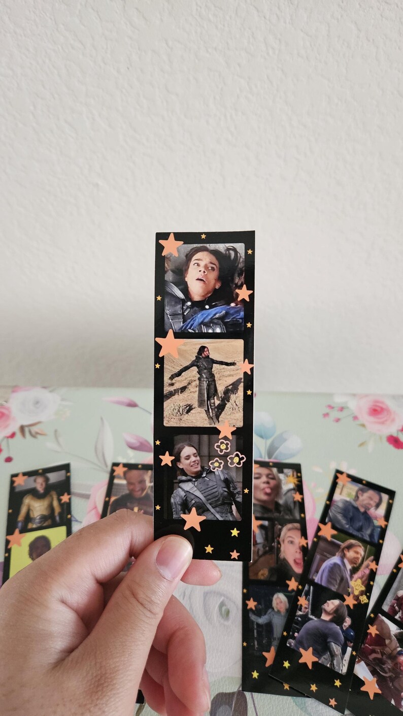 May include: A black bookmark featuring three film strip style images of a person. The bookmark is decorated with orange star and flower stickers. Other bookmarks with similar images are visible.