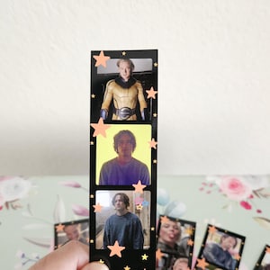May include: Black film strip bookmark with three images of a person, decorated with orange star stickers. The bookmark is held in a hand. Other similar bookmarks are visible in the background.