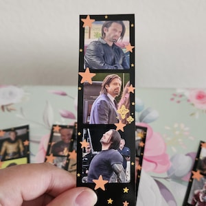 May include: A black bookmark featuring three film strip-style images of a man. The bookmark is decorated with orange star shapes. The images show the man in different outfits and poses. The bookmark is held by a hand.