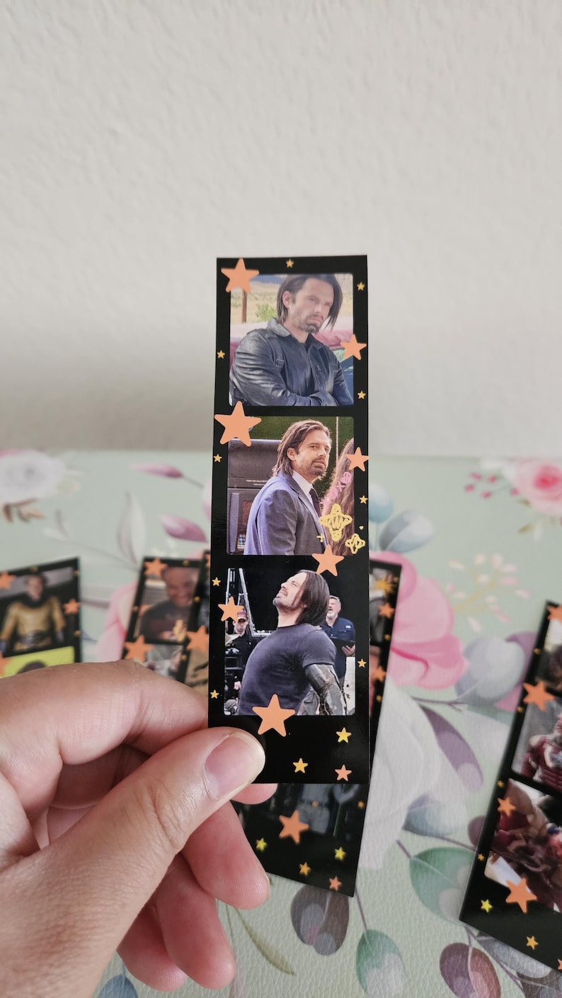 May include: A black bookmark featuring three film strip-style images of a man. The bookmark is decorated with orange star shapes. The images show the man in different outfits and poses. The bookmark is held by a hand.