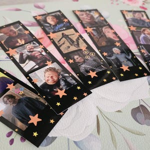 May include: A collection of black film strip-style bookmarks featuring various images of people. Each bookmark has a series of small photo frames with images and is decorated with orange star accents. The bookmarks are arranged on a floral patterned surface.