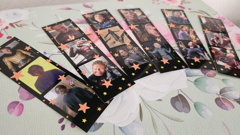 May include: A collection of black film strip-style bookmarks featuring various images of people. Each bookmark has a series of small photo frames with images and is decorated with orange star accents. The bookmarks are arranged on a floral patterned surface.