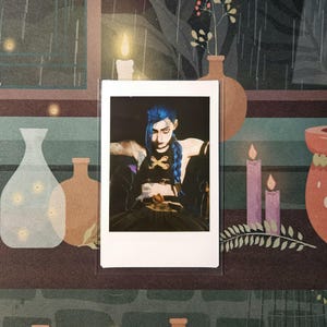 May include: A photograph of a person with blue hair wearing a black outfit and a necklace with a skull and crossbones design. The photo is printed on a white background with a white border.