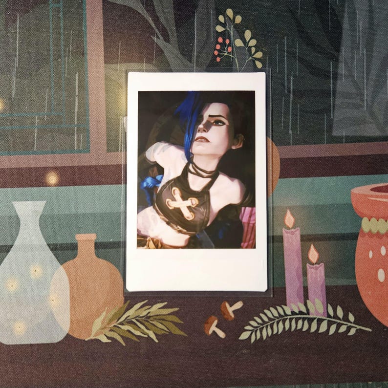 May include: A Polaroid photograph of a woman with blue hair wearing a brown leather top with a cross design. The photo is framed in a white border and is sitting on a table with a dark brown surface.