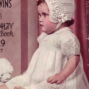 Puede incluir: Una niña pequeña con un vestido y un gorro de ganchillo blanco. El vestido tiene un delicado patrón de encaje y el gorro tiene un lazo en un lado. La bebé está sentada en una superficie roja y sostiene un cordero de peluche blanco.