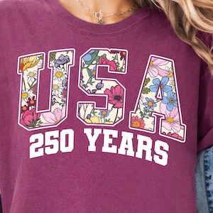 Könnte beinhalten: Beerenfarbenes T-Shirt mit dem weißen Text "USA 250 YEARS". Die Buchstaben "USA" sind mit einem Blumenmuster aus rosa, gelben und blauen Blumen gefüllt. Das Shirt hat hochgekrempelte Ärmel und wird mit einer Jeansjacke getragen.