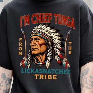 Könnte beinhalten: Ein schwarzes kurzärmeliges T-Shirt mit einer Grafik einer Person, die einen Federschmuck trägt. Der Text lautet I'M CHIEF TUNGA FROM THE LICKASNATCHEE TRIBE, flankiert von zwei Pfeilen neben der zentralen Figur. Das Design verwendet Rot, Blau und Beige.