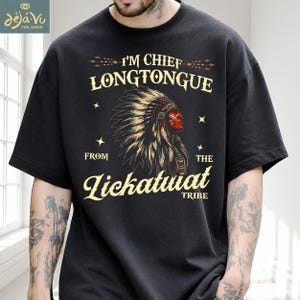 May include: A black short-sleeved t-shirt featuring a graphic of a person wearing a feathered headdress in profile. The text reads I'M CHIEF LONGTONGUE FROM THE LICKATUAT TRIBE in cream-coloured lettering, accented by small stars and arrow motifs.
