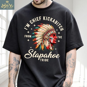 Könnte beinhalten: Ein schwarzes kurzärmeliges T-Shirt mit einer Grafik einer Person, die einen bunten Federschmuck trägt. Der Text I'M CHIEF KICKABITCH FROM THE Slapahoe TRIBE ist kreisförmig um die zentrale Illustration angeordnet.