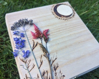 Puede incluir: Una caja de madera con tapa con bisagras, decorada con flores prensadas y un diseño de luna creciente quemado en madera. Las flores son de color azul claro, rosa y verde.