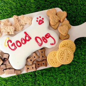 Könnte beinhalten: Ein weißer knochenförmiger Keks mit rotem Zuckerguss, auf dem "Good Dog" steht, auf einem Holzbrett mit verschiedenen Leckerlis für Hunde, darunter herzförmige Kekse, knochenförmige Kekse und kleine quadratische Kekse.