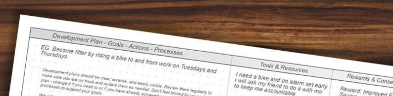 May include: A white printed planner page with the title "Development Plan - Goals - Actions - Processes" on a wooden surface. The text includes "Become fitter by riding a bike" and "Tools & Resources".