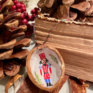Könnte beinhalten: Ein handbemaltes Nussknacker-Ornament in einer halbierten Walnussschale. Der Nussknacker trägt eine rot-blaue Uniform mit einem goldenen Hut und weißen Verzierungen. Das Ornament ist mit goldenem Glitzer und grünen Tannenzweigen verziert.