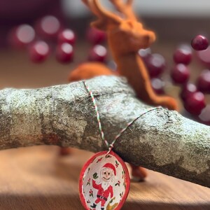 Könnte beinhalten: Handbemalter Weihnachtsschmuck mit dem Weihnachtsmann, mit rotem Rand und weißem Innenraum. Der Schmuck hängt an einem Ast, mit einer verschwommenen Rentierfigur im Hintergrund. Der Schmuck hat einen Durchmesser von etwa 5 cm.