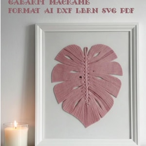 May include: A framed pink macrame leaf art piece. The leaf is centered in a white frame, with the text "MACRAME TEMPLATE GABARIT MACRAME FORMAT AI DXF LBRN SVG PDF" above the frame. A lit candle sits to the left.