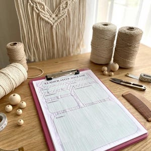 May include: A macrame project planning setup on a wooden surface. Includes a pink clipboard with a planning sheet, spools of beige and brown yarn, wooden beads, scissors, a comb, and a measuring tape. A macrame wall hanging is in the background.
