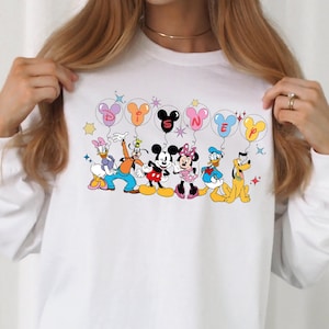 May include: White long-sleeve shirt with a graphic print of Mickey Mouse and friends holding balloons that spell out "DISNEY".