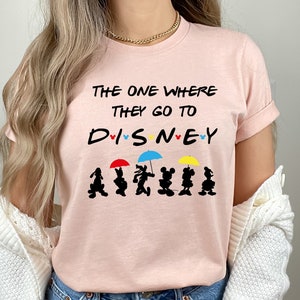 May include: A pink t-shirt with the text "The One Where They Go To Disney" and a line of black silhouettes of Mickey Mouse and friends holding umbrellas.