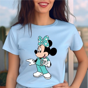 May include: A light blue t-shirt with a cartoon image of Minnie Mouse dressed as a doctor. She is wearing a turquoise scrub top, a stethoscope, and a white polka dot bow.