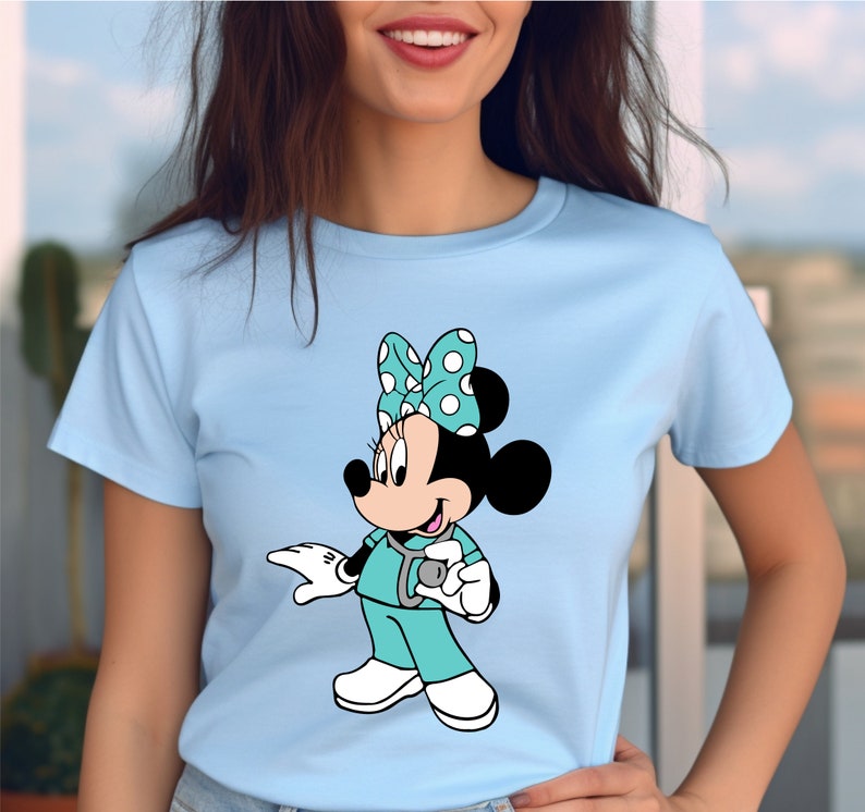 May include: A light blue t-shirt with a cartoon image of Minnie Mouse dressed as a doctor. She is wearing a turquoise scrub top, a stethoscope, and a white polka dot bow.