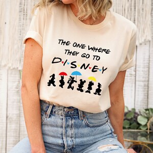 May include: A beige t-shirt with the text "The One Where They Go To Disney" and a line of Disney characters holding umbrellas.