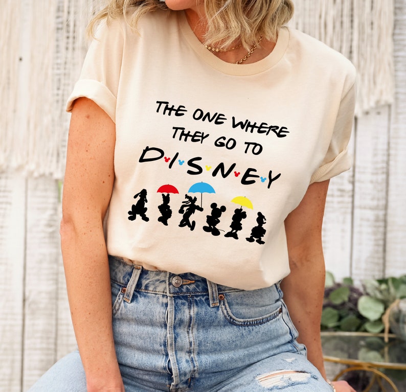 May include: A beige t-shirt with the text "The One Where They Go To Disney" and a line of Disney characters holding umbrellas.