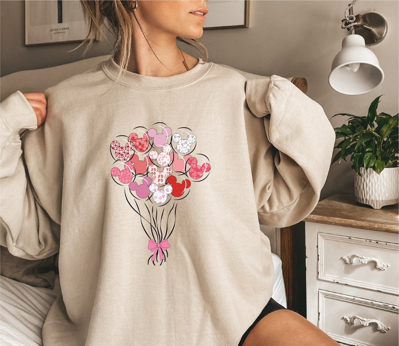 May include: A beige sweatshirt with a graphic of pink and red Mickey Mouse balloons. The balloons are tied together with a pink bow.