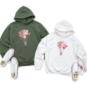 May include: Two hooded sweatshirts with a graphic of a bouquet of Mickey Mouse balloons. The sweatshirt on the left is olive green and the sweatshirt on the right is white.