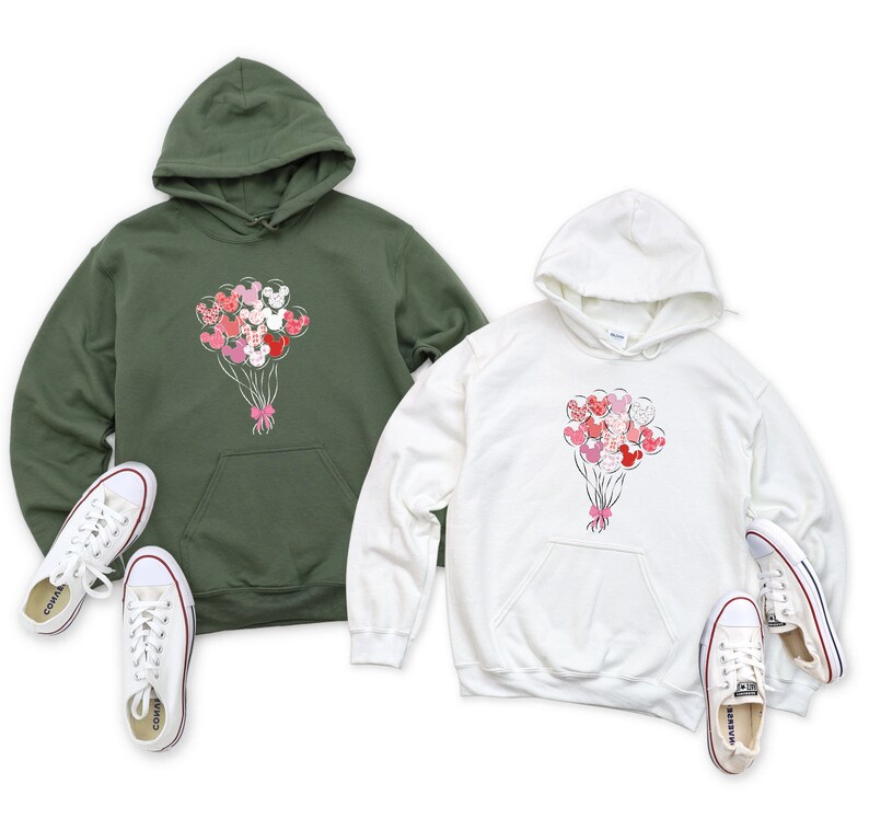 May include: Two hooded sweatshirts with a graphic of a bouquet of Mickey Mouse balloons. The sweatshirt on the left is olive green and the sweatshirt on the right is white.