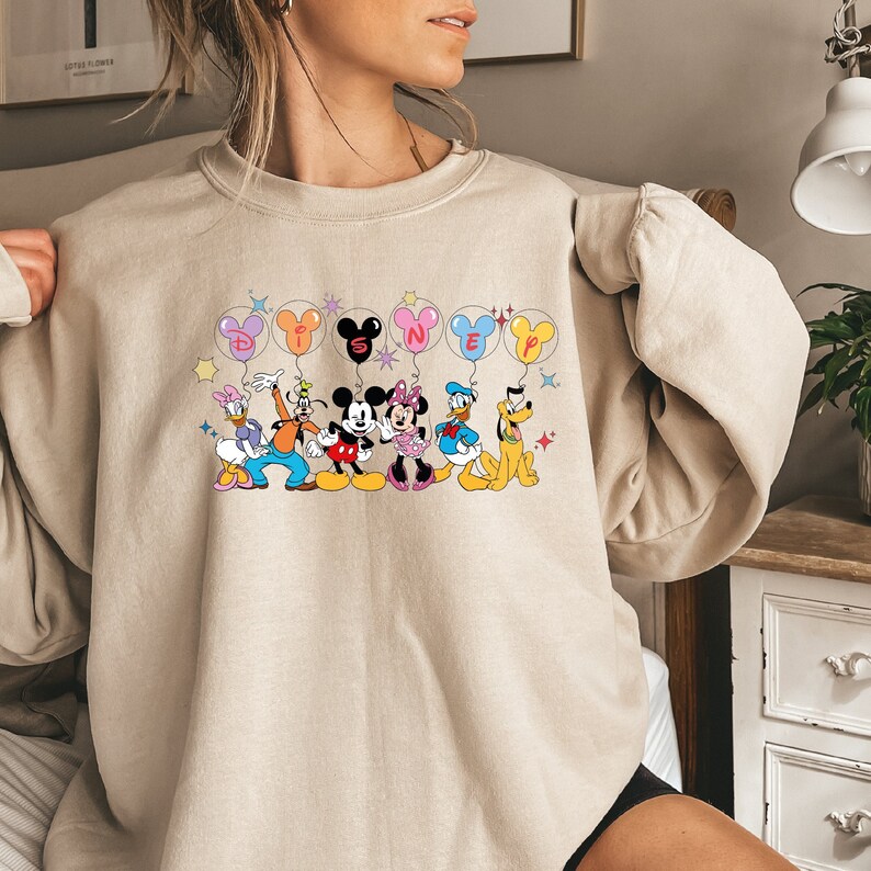 May include: A beige sweatshirt with a graphic print of Mickey Mouse, Minnie Mouse, Donald Duck, Goofy, and Pluto holding balloons that spell out "DISNEY".