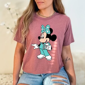 May include: A mauve cropped t-shirt with a graphic of a cartoon Minnie Mouse dressed as a nurse. Minnie Mouse is wearing a teal scrub top, a white stethoscope, and a blue and white polka dot bow.