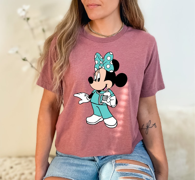 May include: A mauve cropped t-shirt with a graphic of a cartoon Minnie Mouse dressed as a nurse. Minnie Mouse is wearing a teal scrub top, a white stethoscope, and a blue and white polka dot bow.