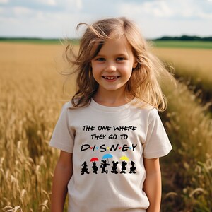 May include: A young girl wearing a white t-shirt with a black graphic that says "The one where they go to DISNEY" with Mickey Mouse characters and umbrellas. The girl is standing in a field of tall grass.
