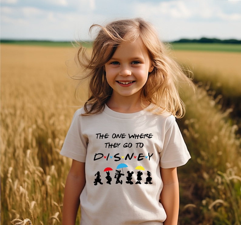 May include: A young girl wearing a white t-shirt with a black graphic that says "The one where they go to DISNEY" with Mickey Mouse characters and umbrellas. The girl is standing in a field of tall grass.