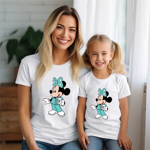 May include: A white t-shirt with a cartoon image of Minnie Mouse dressed as a doctor. The shirt has a teal blue bow with white polka dots. The image is repeated on a smaller white t-shirt.