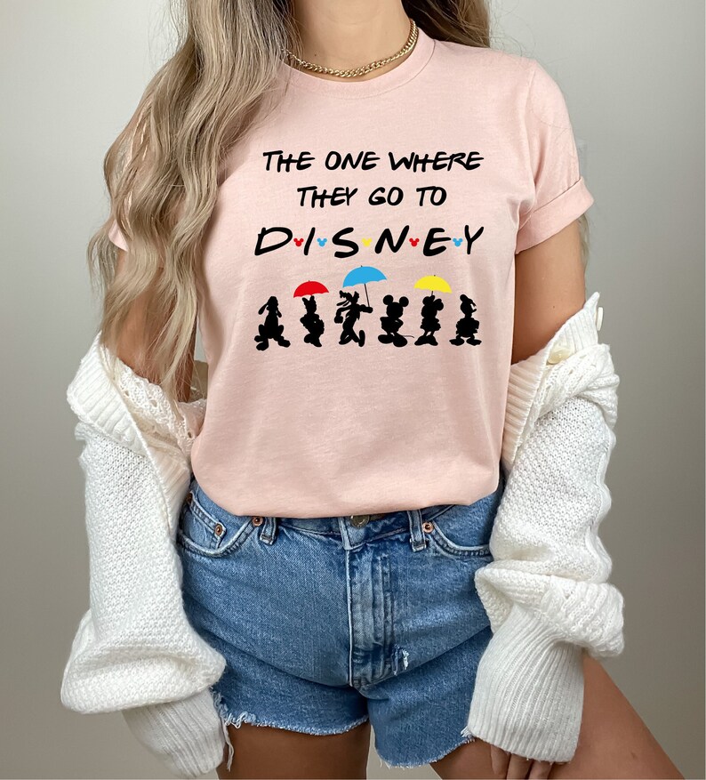 May include: A pink t-shirt with the text "The One Where They Go To Disney" and a line of black silhouettes of Mickey Mouse and friends holding umbrellas.