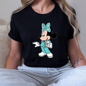 May include: Black t-shirt with a graphic of a cartoon Minnie Mouse dressed as a nurse. Minnie Mouse is wearing a turquoise scrub top, a stethoscope, and a red and white polka dot bow.