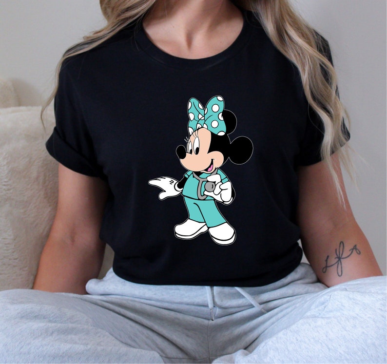 May include: Black t-shirt with a graphic of a cartoon Minnie Mouse dressed as a nurse. Minnie Mouse is wearing a turquoise scrub top, a stethoscope, and a red and white polka dot bow.