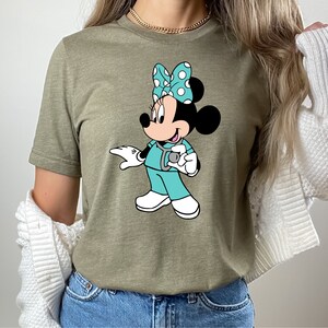 May include: A green t-shirt with a cartoon image of Minnie Mouse dressed as a nurse. Minnie is wearing a blue scrub top, a stethoscope, and a white bow with blue polka dots.