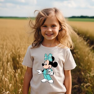 May include: A young girl wearing a white t-shirt with a cartoon image of Minnie Mouse dressed as a doctor. Minnie Mouse is wearing a turquoise scrub top with white polka dots and a white bow with blue polka dots. She is holding a stethoscope in her right hand.