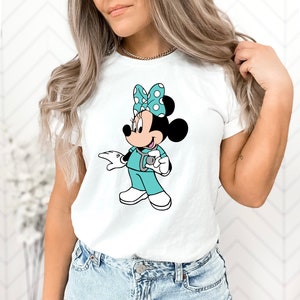 May include: White t-shirt with a cartoon image of Minnie Mouse dressed as a nurse. Minnie Mouse is wearing a teal scrub top, a stethoscope, and a white polka dot bow.