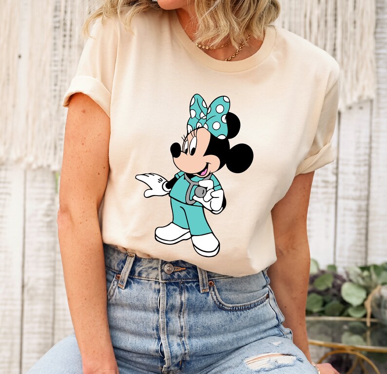 May include: A beige t-shirt with a graphic of Minnie Mouse dressed as a nurse. Minnie is wearing a turquoise scrub top, a stethoscope, and a white bow with black polka dots.