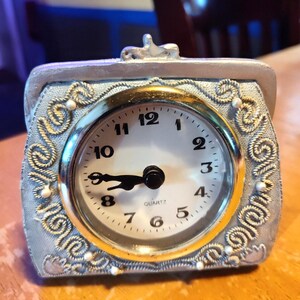 May include: A silver-colored quartz clock in a purse-shaped design. The clock face is white with black numerals and hands, and a gold-colored bezel. The clock is decorated with ornate silver scrollwork and small white beads.